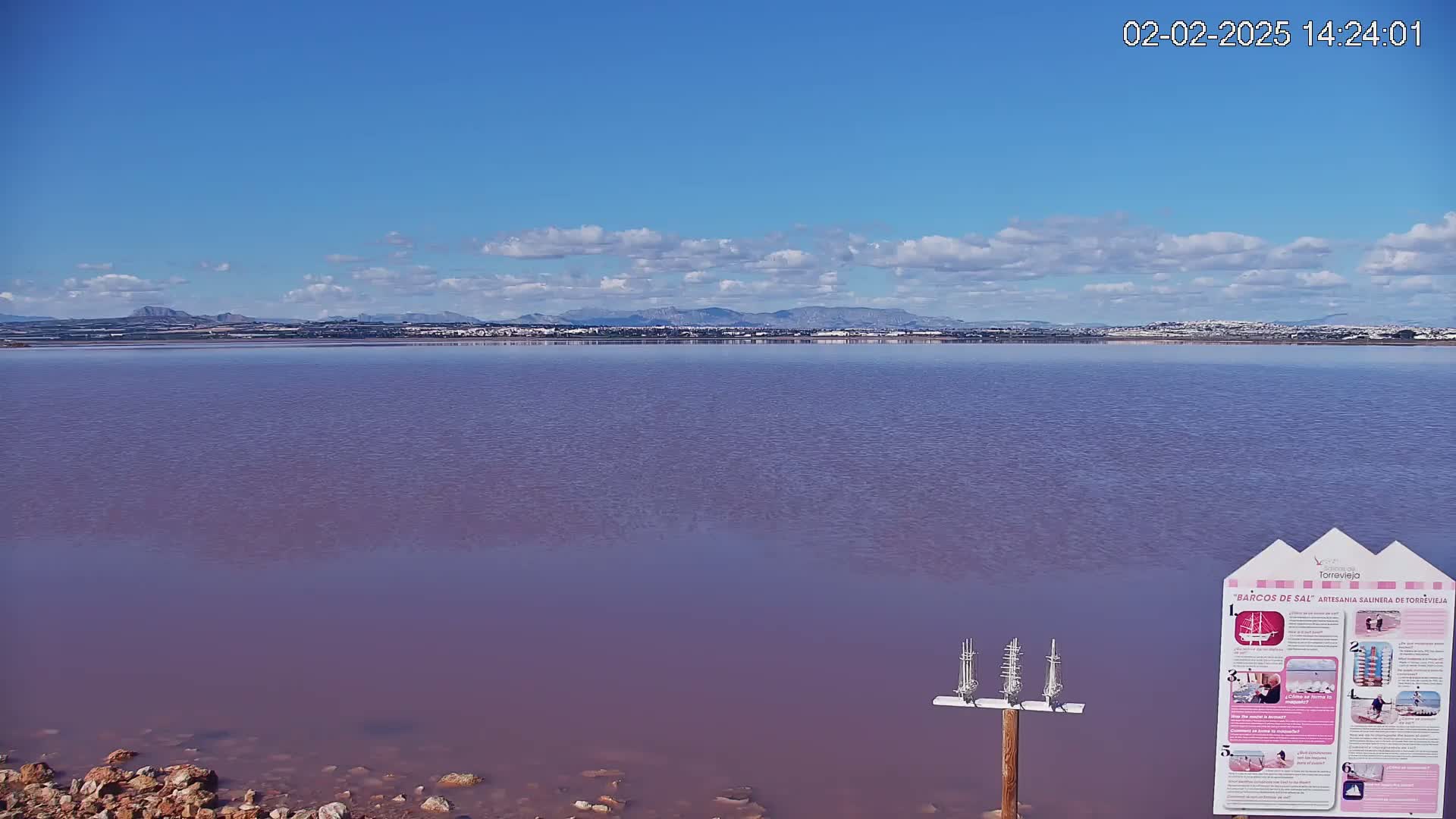 A calm, pinkish lake stretches to a distant shoreline under a mostly clear blue sky with a few scattered clouds.