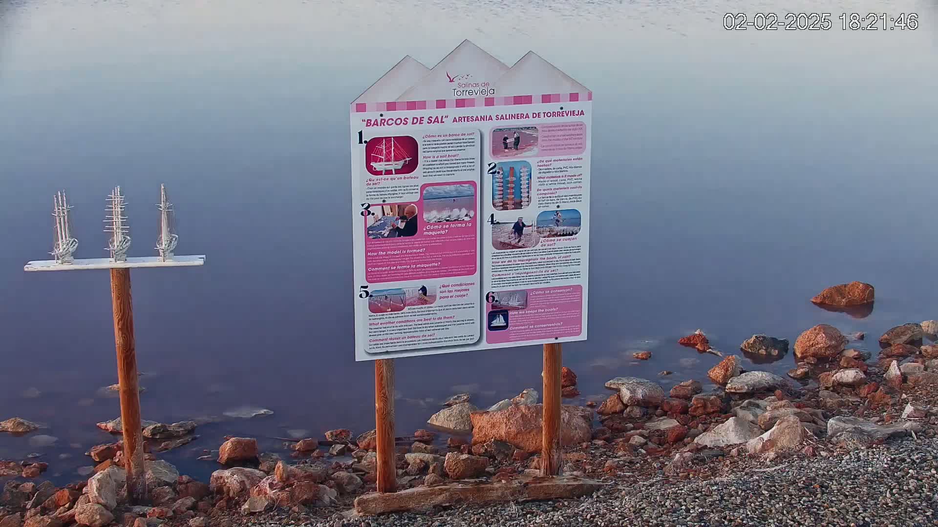 A calm, purplish-blue body of water laps a rocky shore where a wooden post holds three small model sailboats, next to a sign detailing their creation.