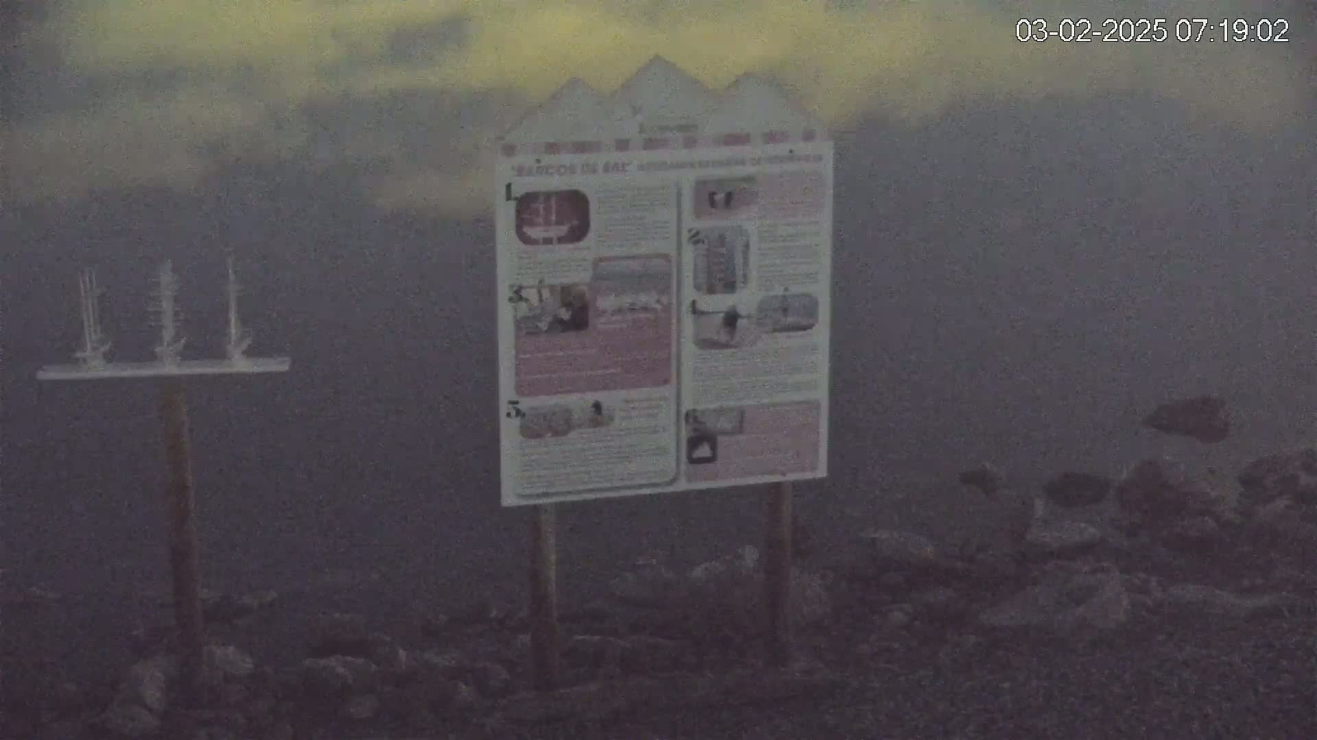 A sign stands on a rocky ground under an overcast sky, with a small model of ships on a post in the background.