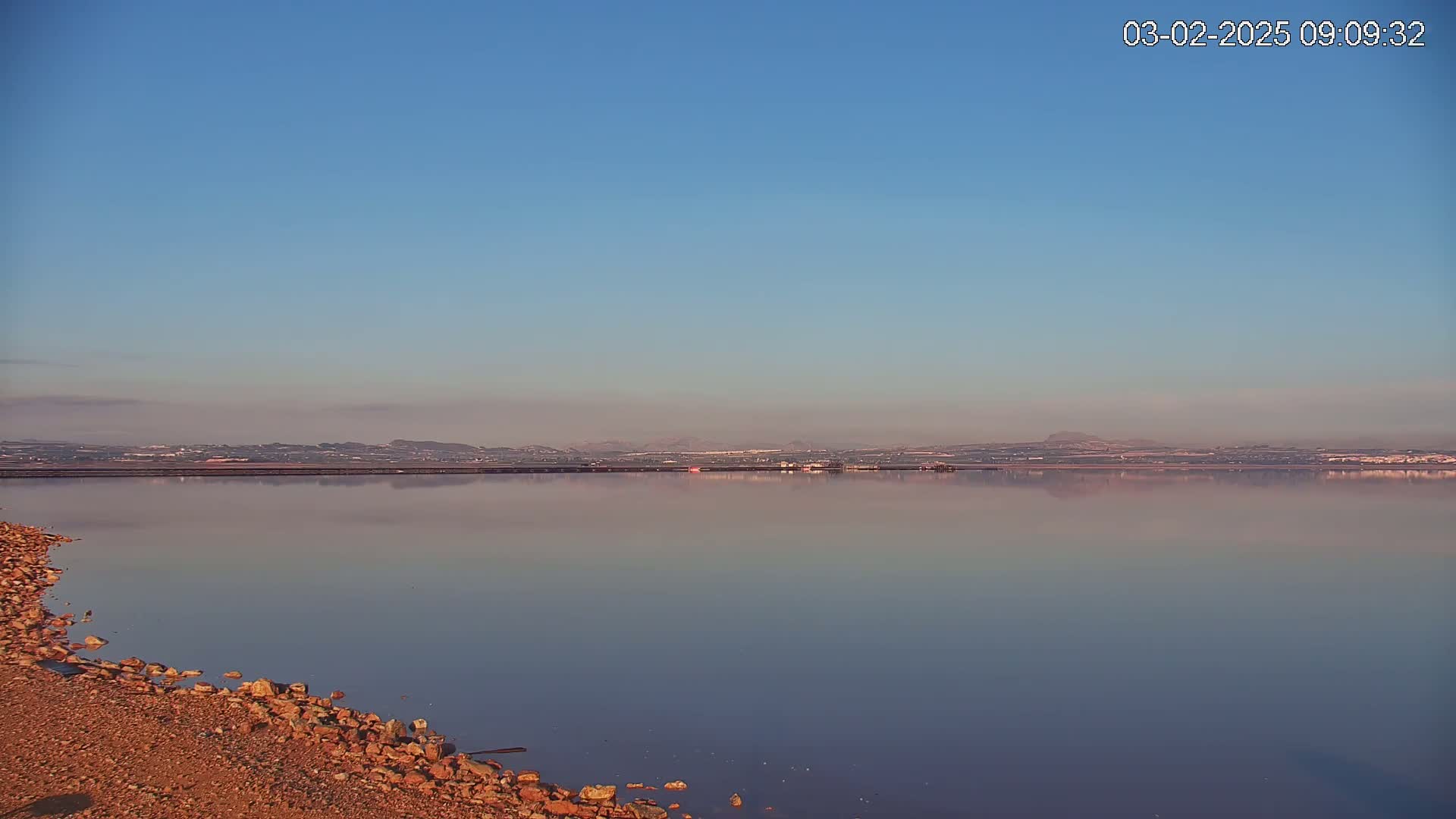 A calm, pink-hued body of water reflects a distant, hazy landscape under a clear blue sky.