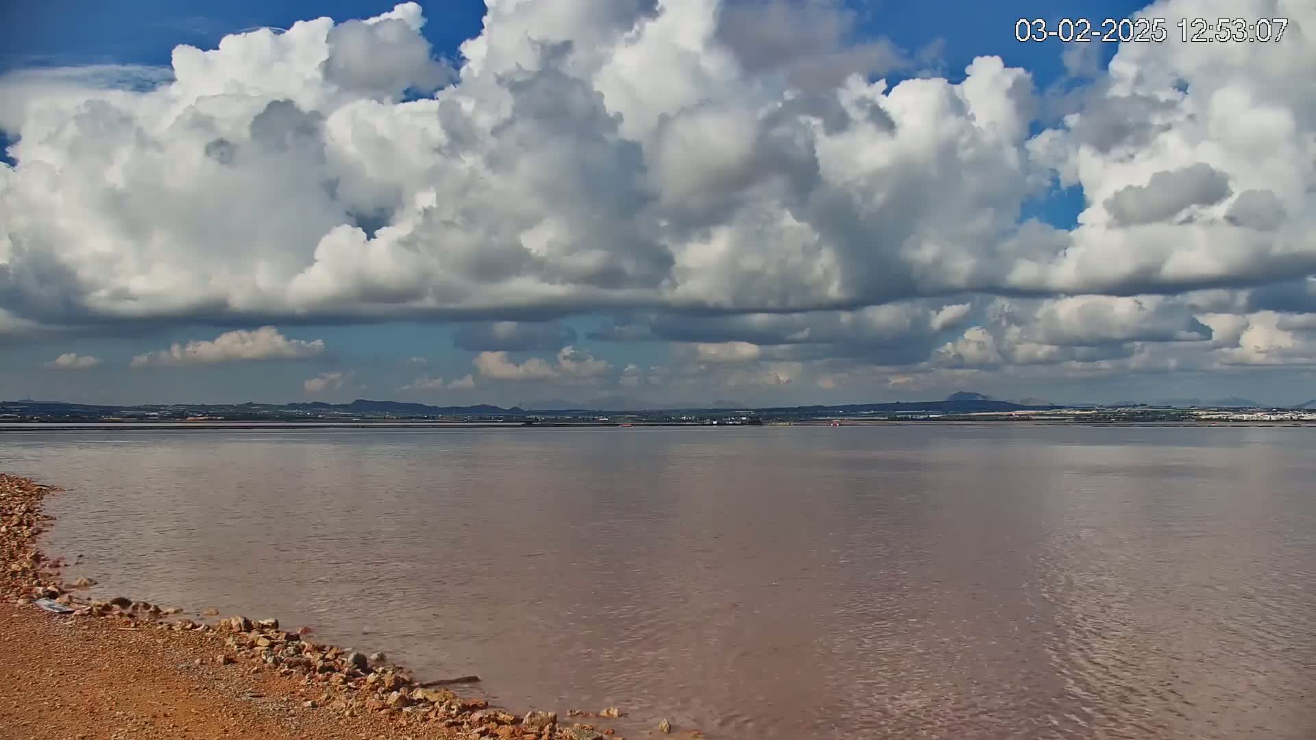 A mostly cloudy sky over a calm body of pinkish-brown water with a rocky shoreline in the foreground and a distant landmass on the horizon.