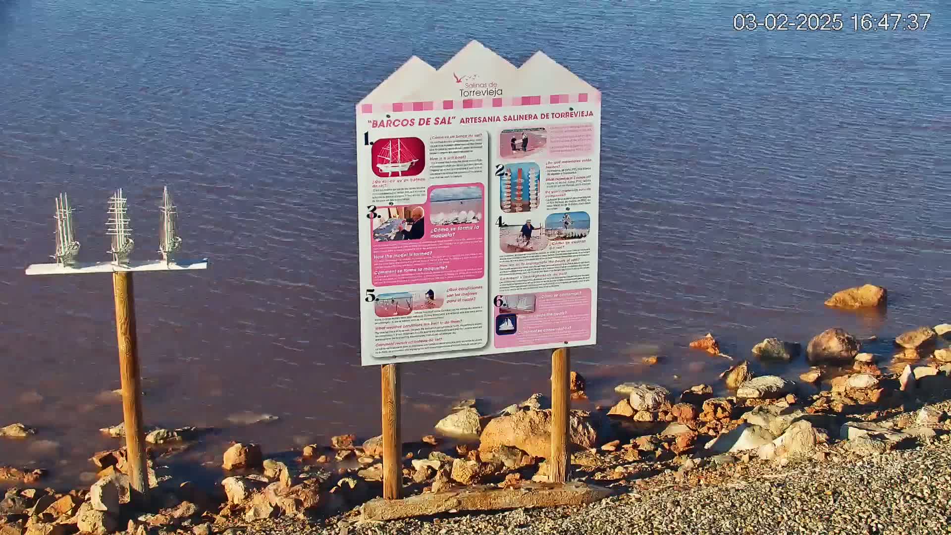A calm body of pinkish-purple water laps a rocky shore near an informational sign and a display of three small model sailboats.