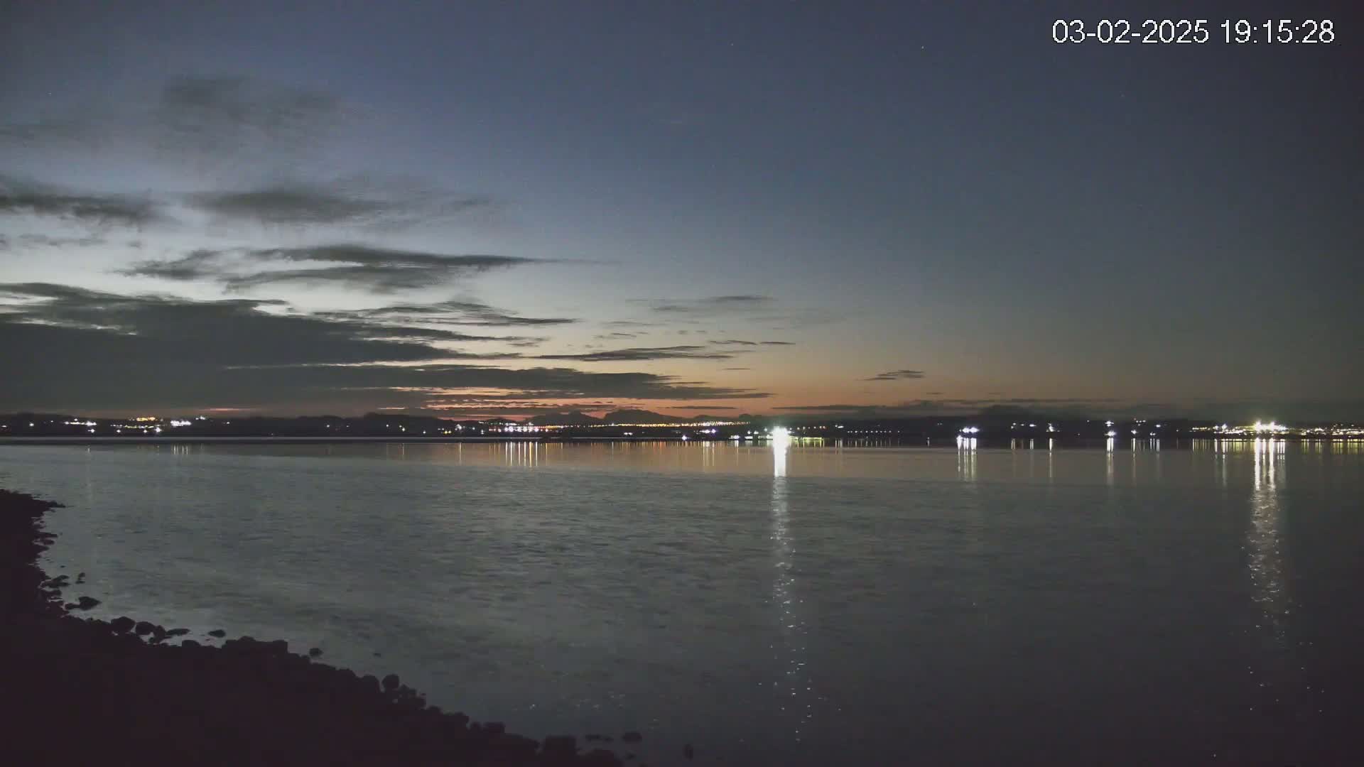 A calm body of water reflects the lights of a distant city skyline under a twilight sky with thin clouds.