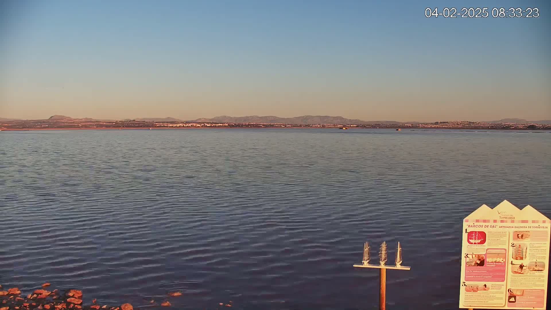 A calm body of water stretches to a distant shoreline under a clear, pale blue sky, with a small sign and model boats visible in the foreground.