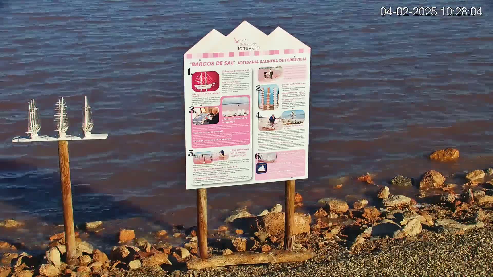 A wooden sign with pictures explaining a salt-making process stands near calm, pinkish-brown water lapping a rocky shore; three small model sailboats are mounted on a post behind the sign.