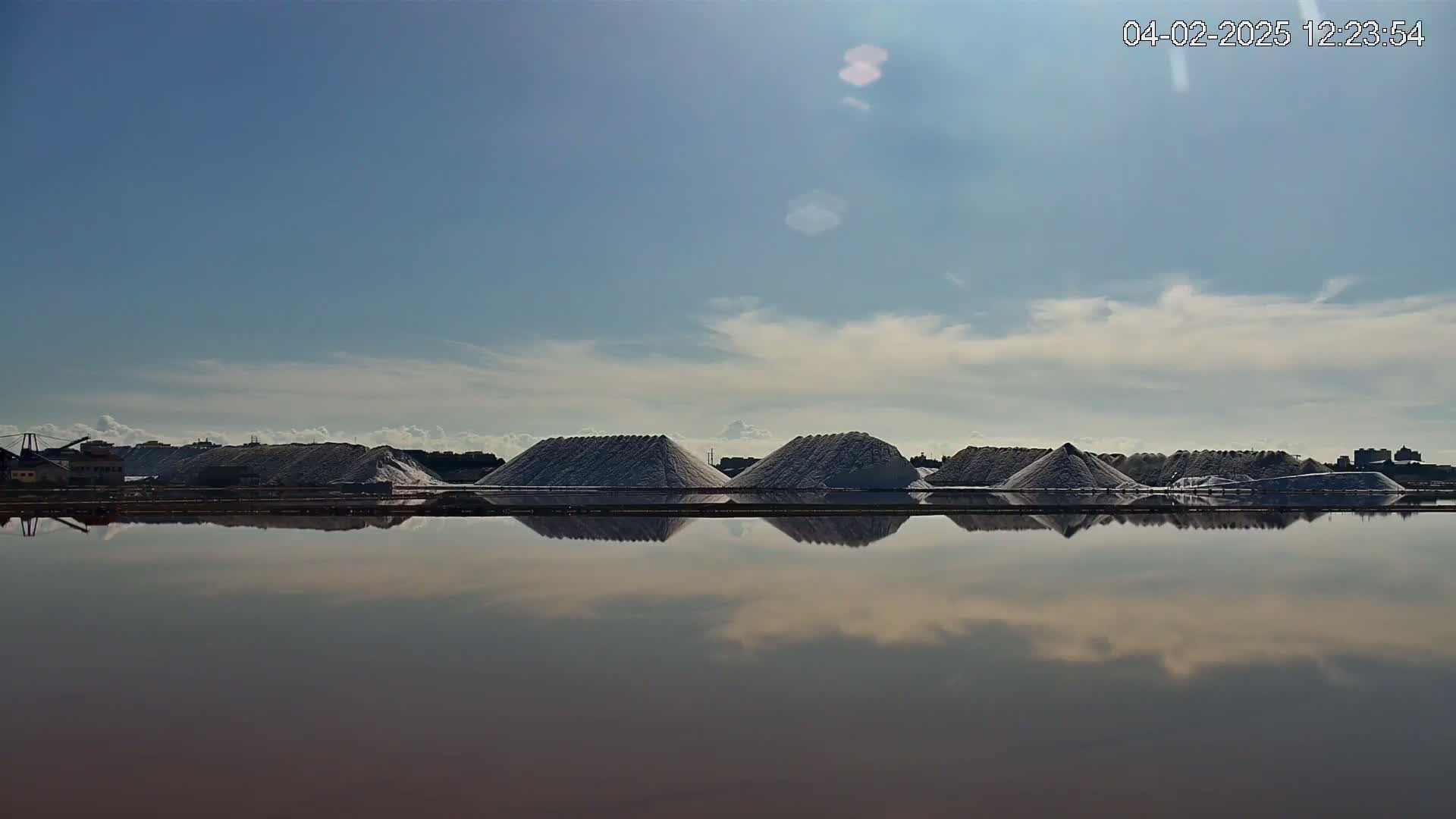Several large piles of salt are reflected in a calm body of water under a mostly clear, bright sky.