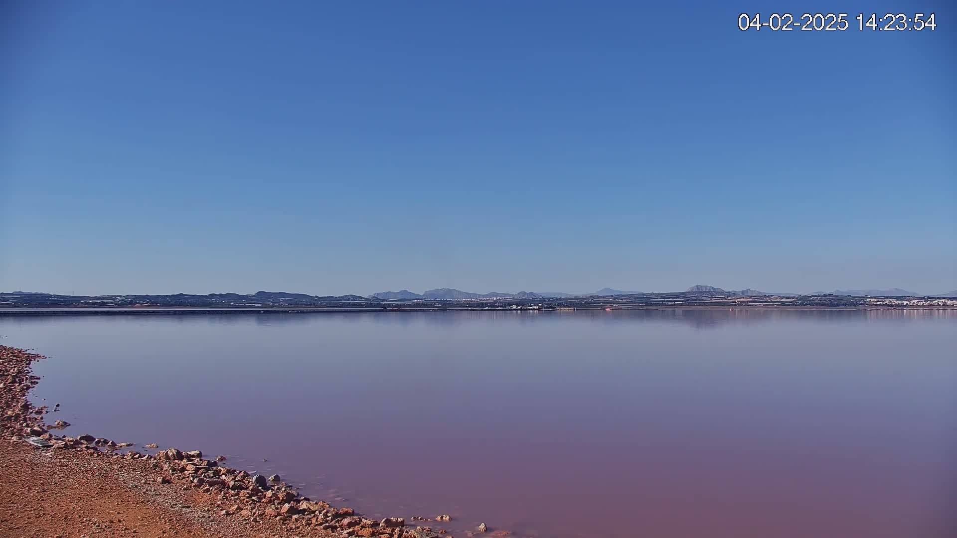 A pinkish lake with a rocky shore stretches to a distant mountain range under a clear blue sky.
