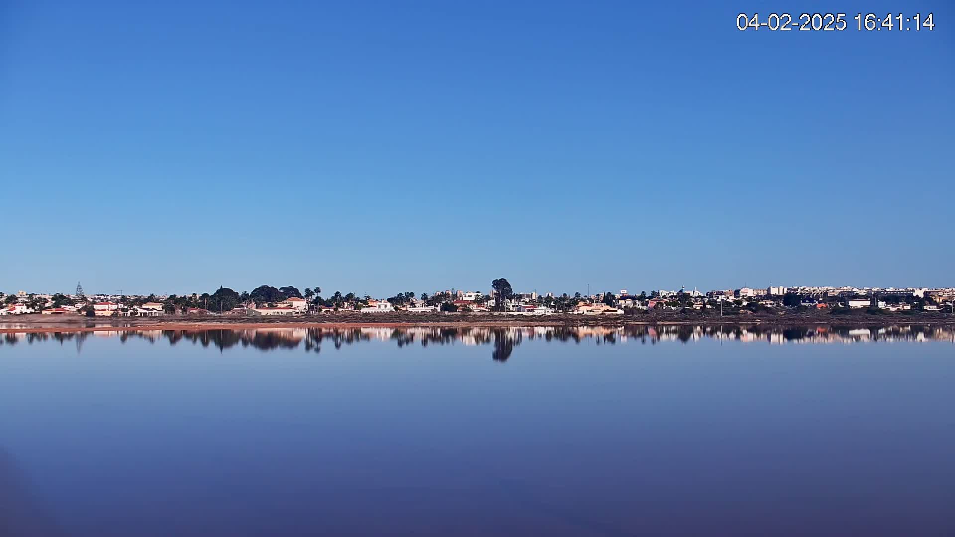 A calm body of water reflects a row of houses and buildings under a clear blue sky.