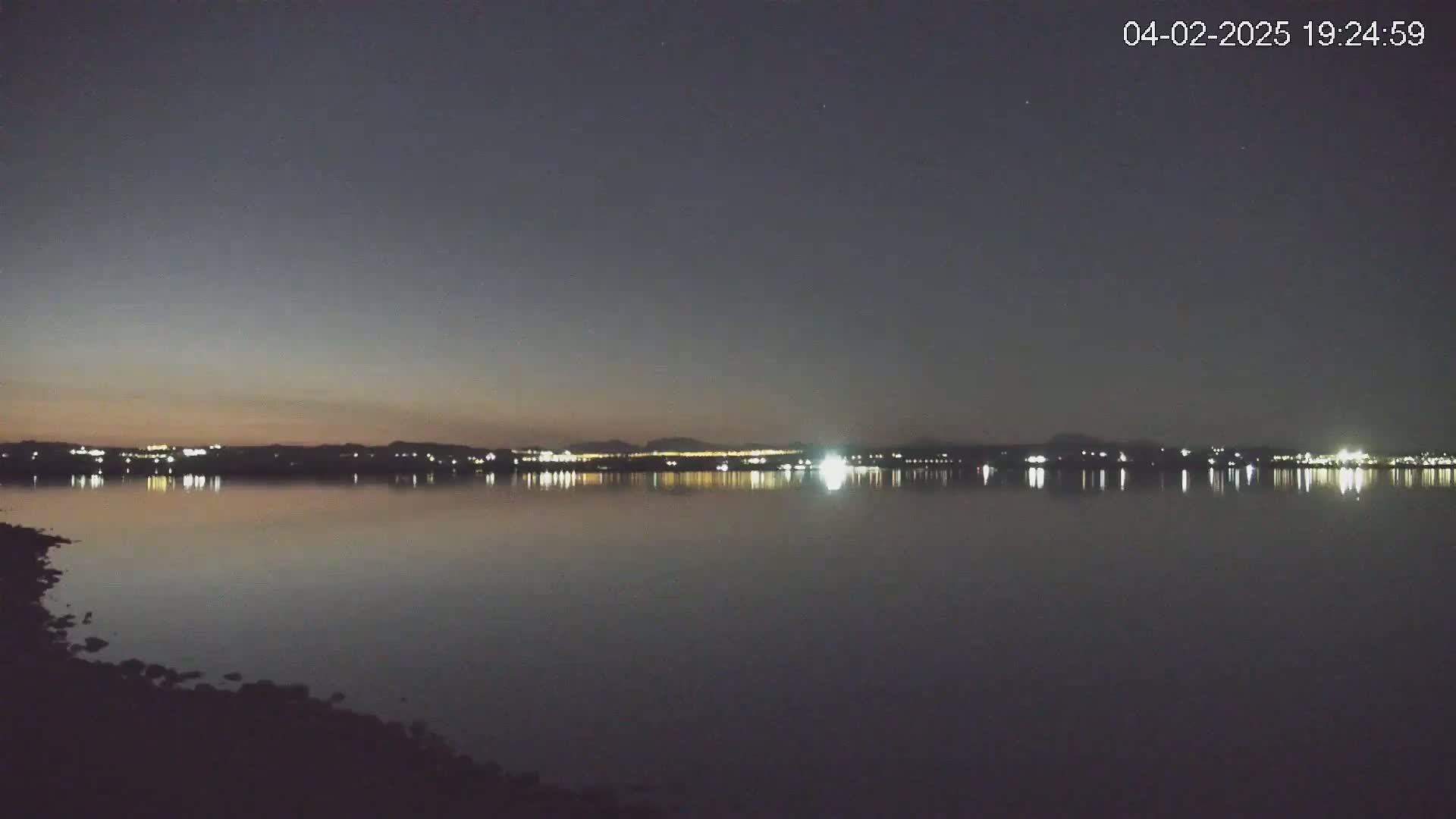 A calm body of water reflects the lights of a distant city skyline under a dark, twilight sky.