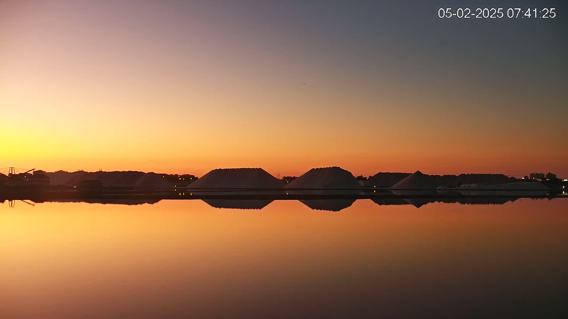 Several large piles of salt are reflected in a calm body of water at sunset.