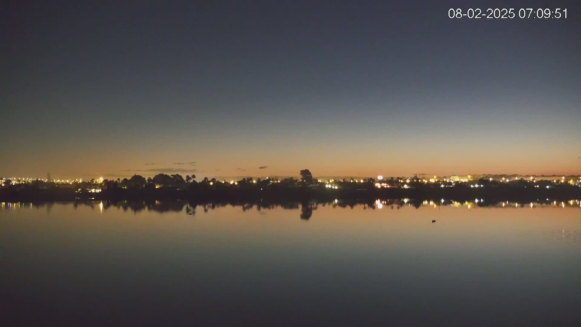 A calm body of water reflects the lights of a city skyline at twilight under a clear, dark sky.