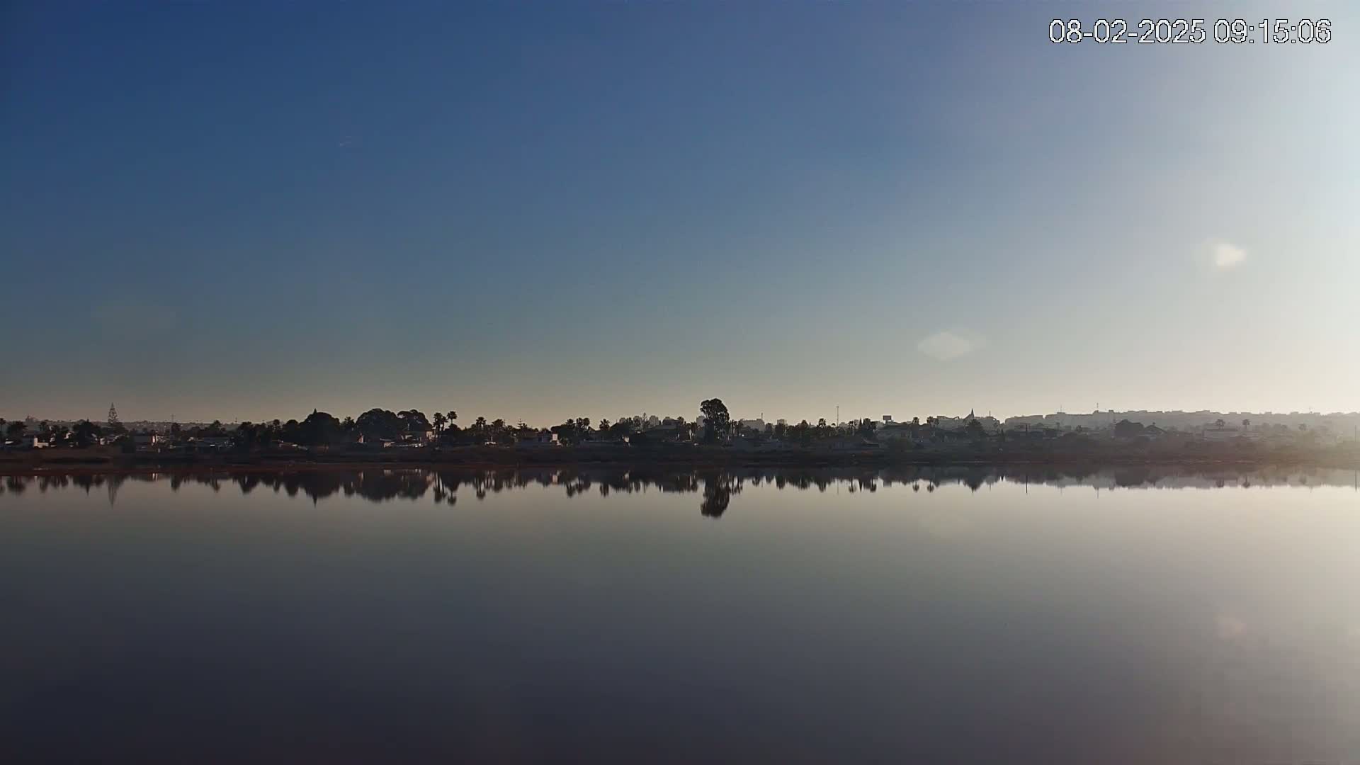 A calm body of water reflects a distant shoreline of houses and trees under a clear, bright sky.