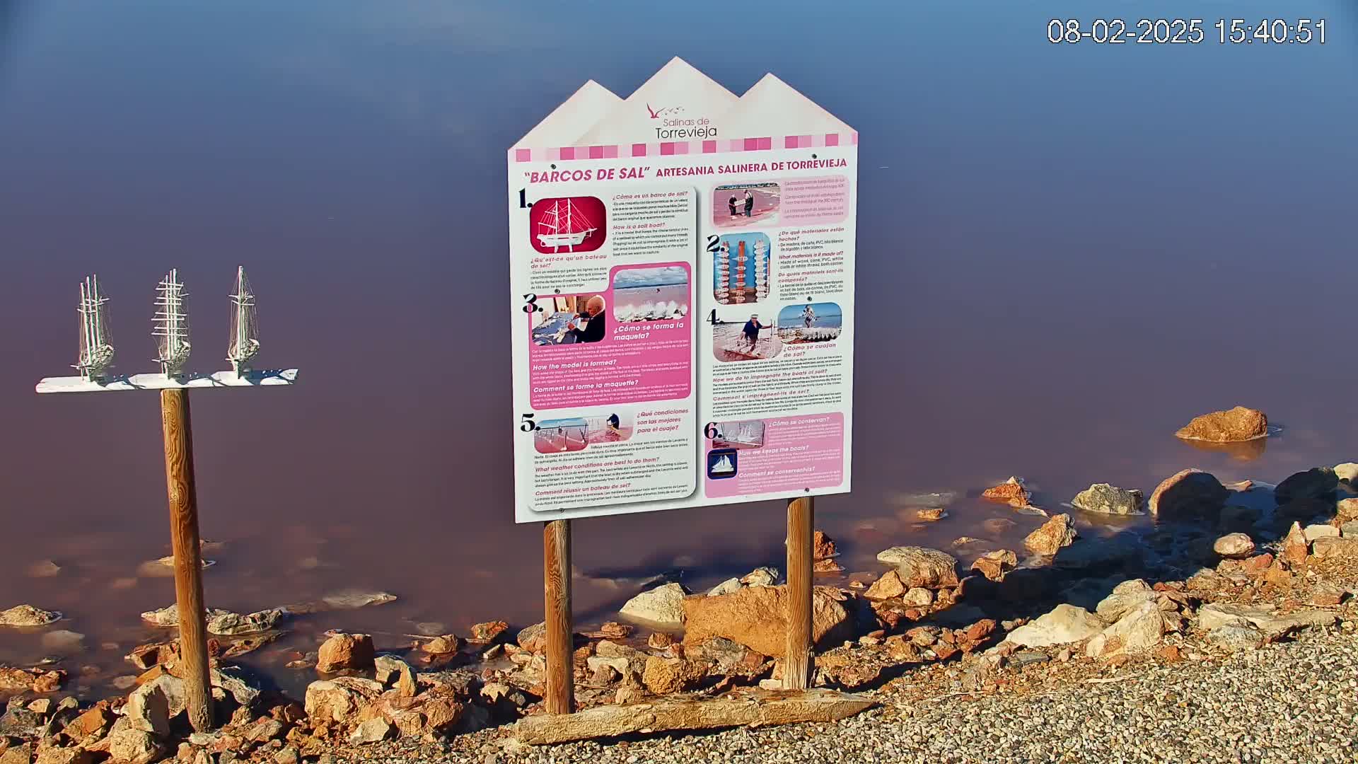 Three miniature model sailing ships stand on a post next to an informational sign by a pinkish-purple body of water with rocks along the shore.