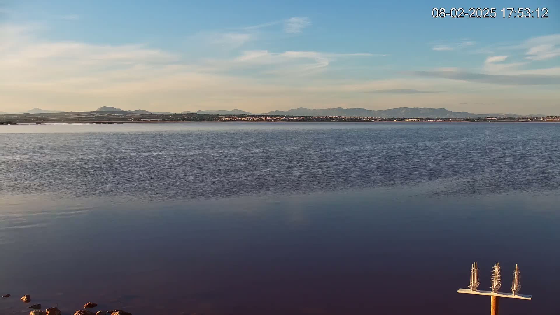 A calm body of water reflects a distant mountain range under a mostly clear sky.
