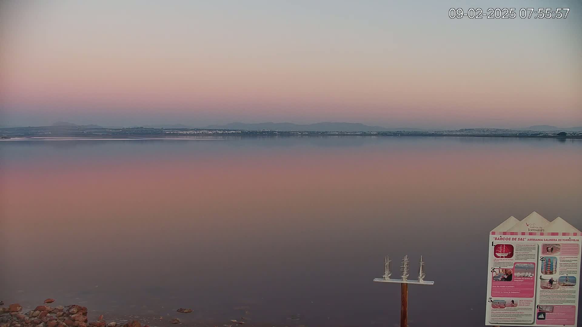 A calm body of pink-tinged water reflects a pale sunrise sky over a distant shoreline.