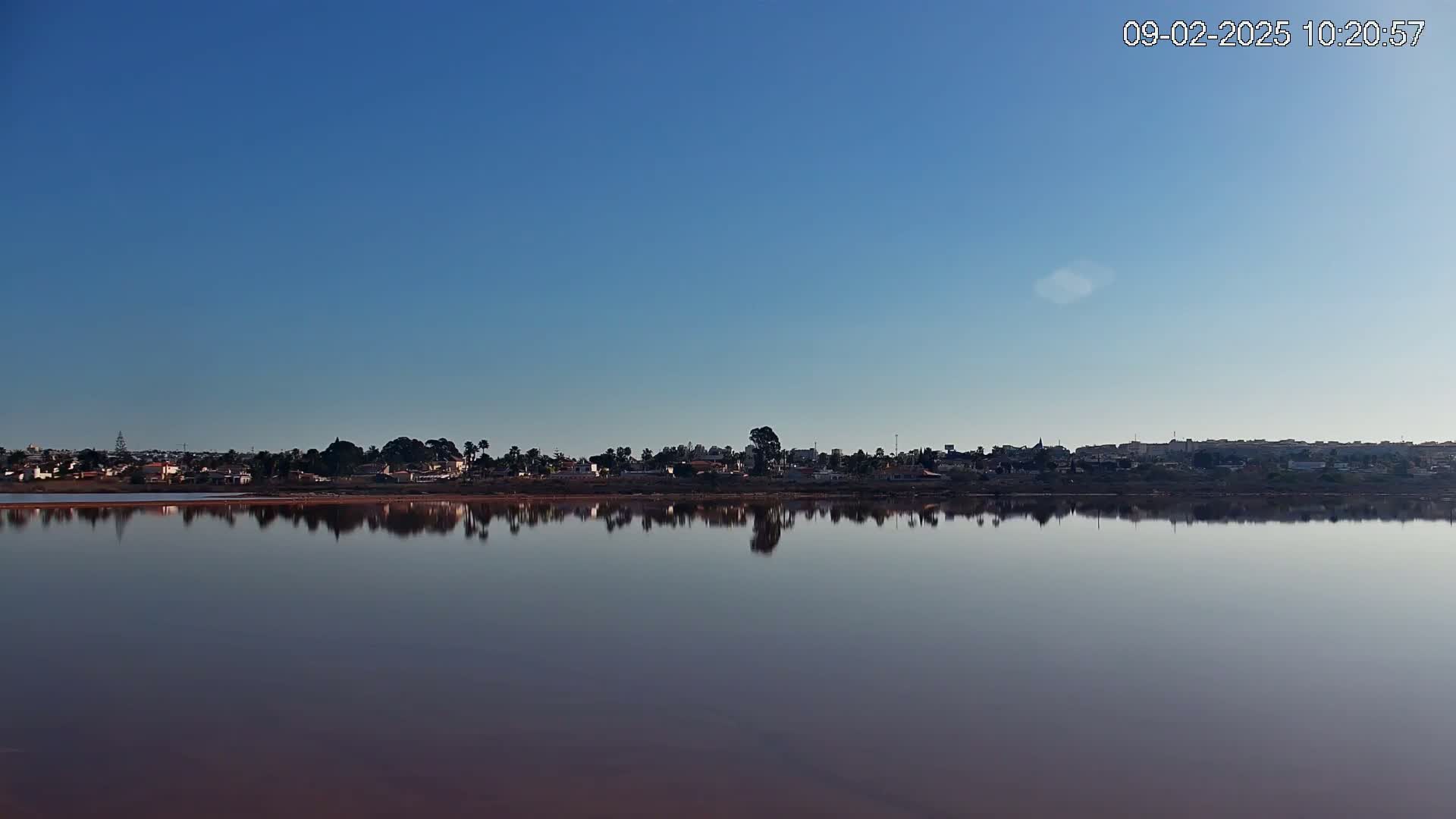 A calm, pink-hued lake reflects a distant town under a clear blue sky.