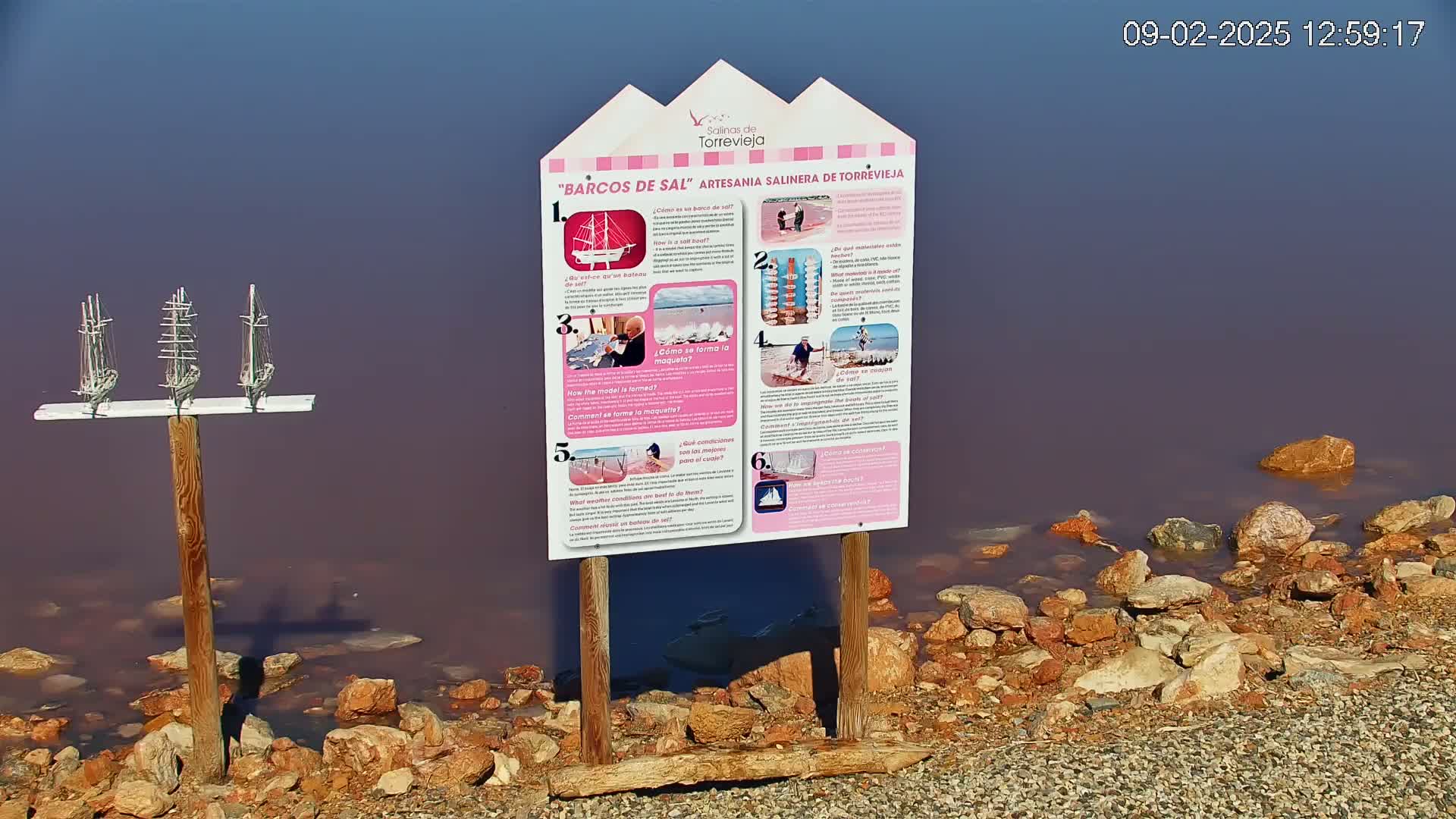 A pink-hued body of water is edged with reddish-brown rocks, and a wooden post displays three miniature ships and a nearby informational sign.