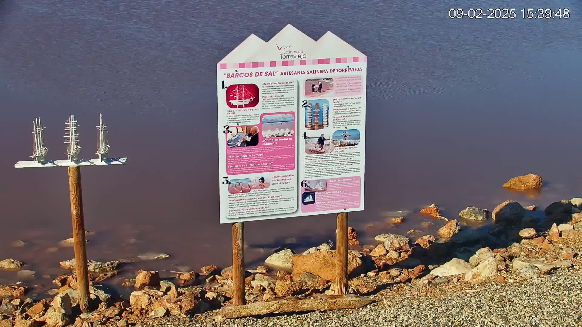 Three miniature model sailboats are mounted on a post near a pink-hued body of water, next to a sign with images and text, and a rocky shoreline.