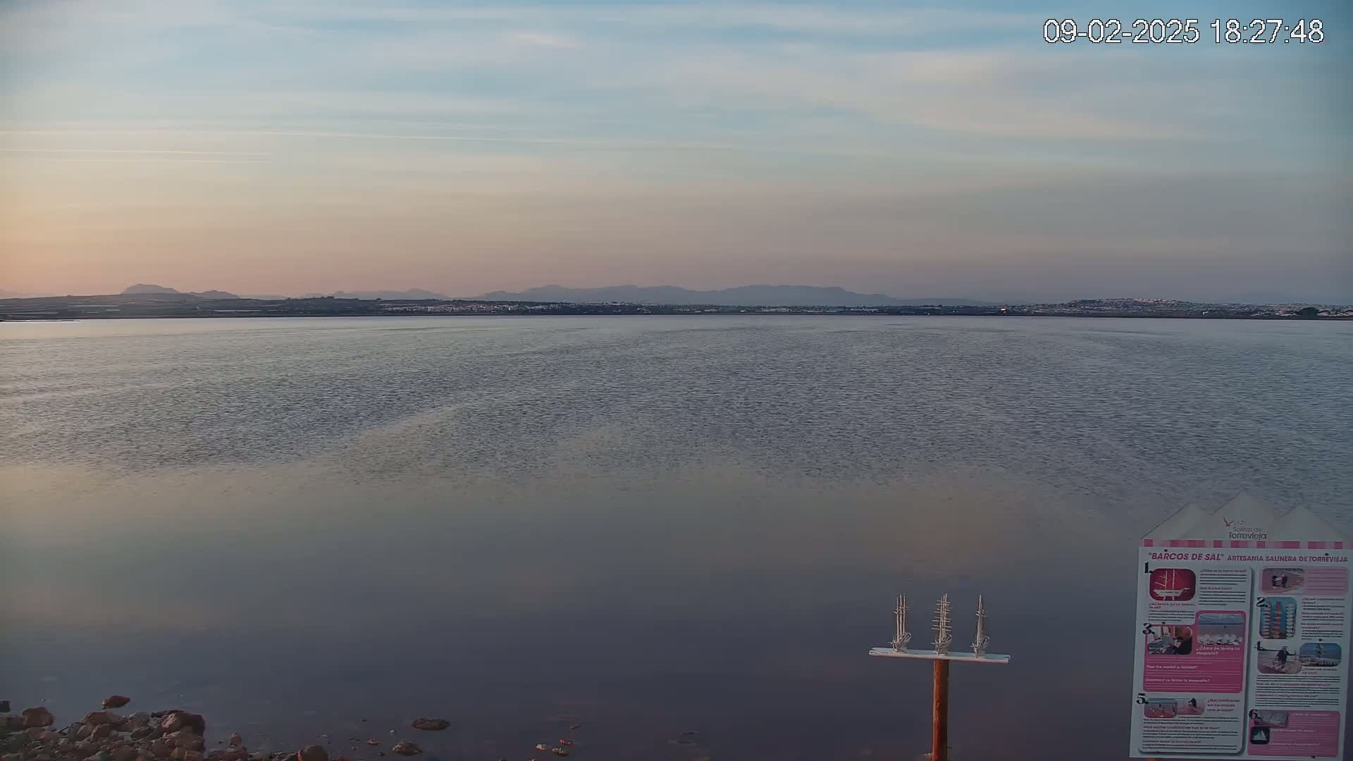 A calm body of water stretches to a distant shoreline under a pale, pinkish sunset sky.