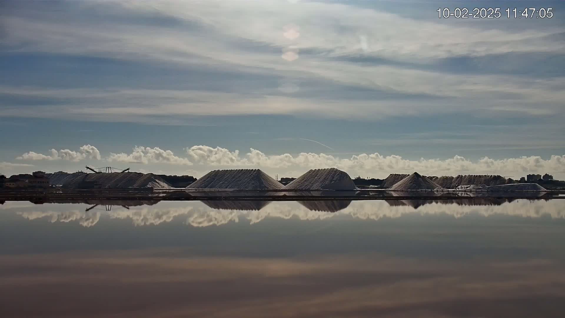 A calm body of water reflects a line of salt piles under a mostly clear sky with scattered cumulus clouds.