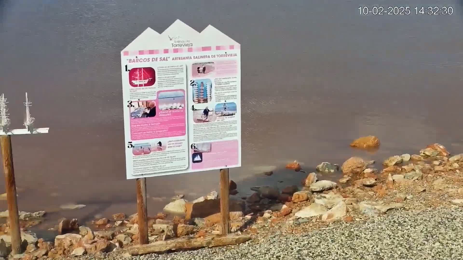 A pink-hued body of water laps against a rocky shore, with an informational sign and small model ships nearby.