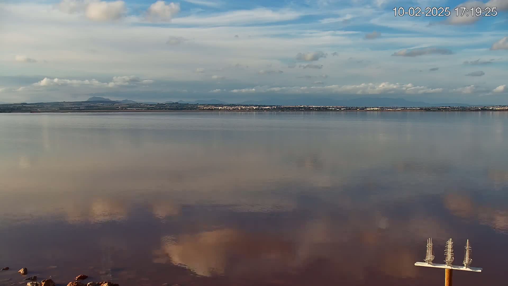 A calm, pinkish-hued body of water reflects a partly cloudy sky and distant land with low hills under a mostly clear sky.