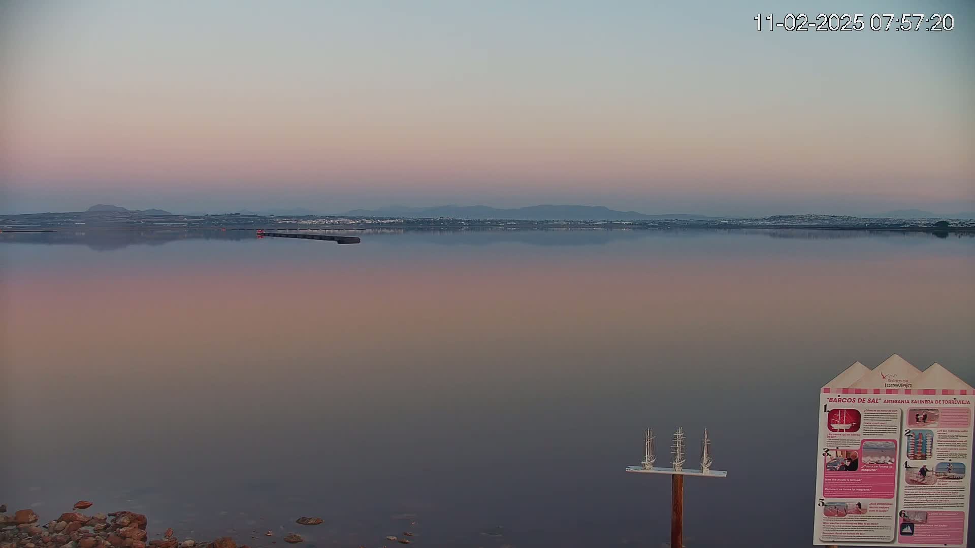 A calm, pink-hued body of water reflects a hazy, distant shoreline under a pale sunrise.