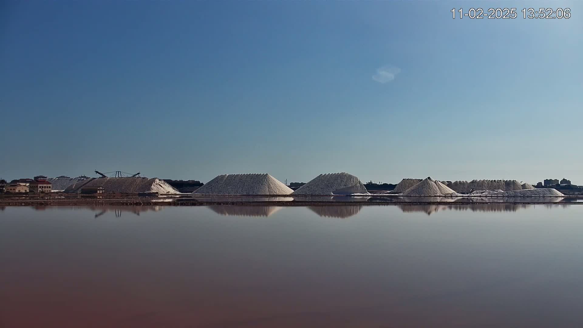 Under a clear blue sky, several large piles of white salt are reflected in a calm, pinkish body of water.