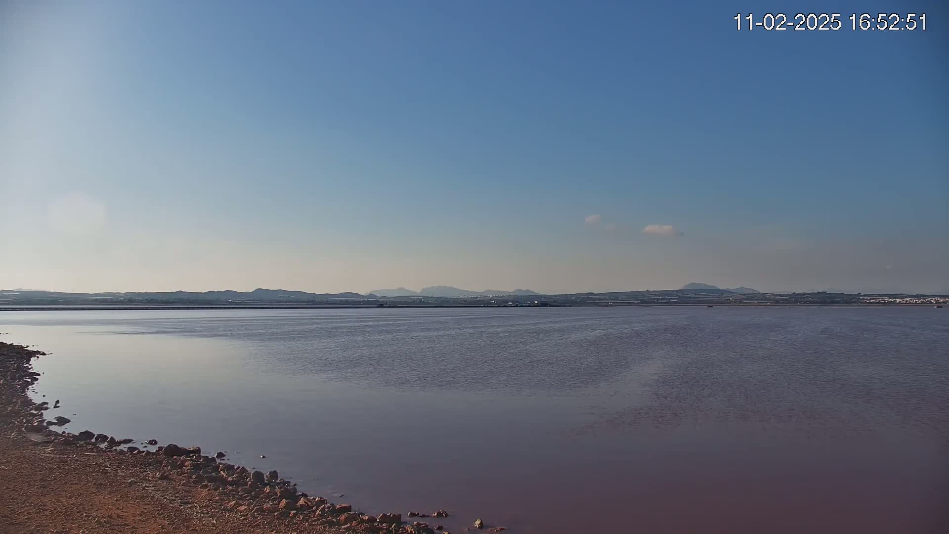 A calm, pinkish body of water stretches towards a distant, low-lying landmass under a clear blue sky.