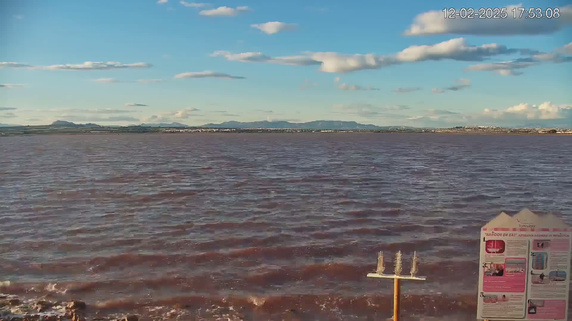 A pinkish-red body of water with small waves stretches to a distant mountain range under a partly cloudy sky.