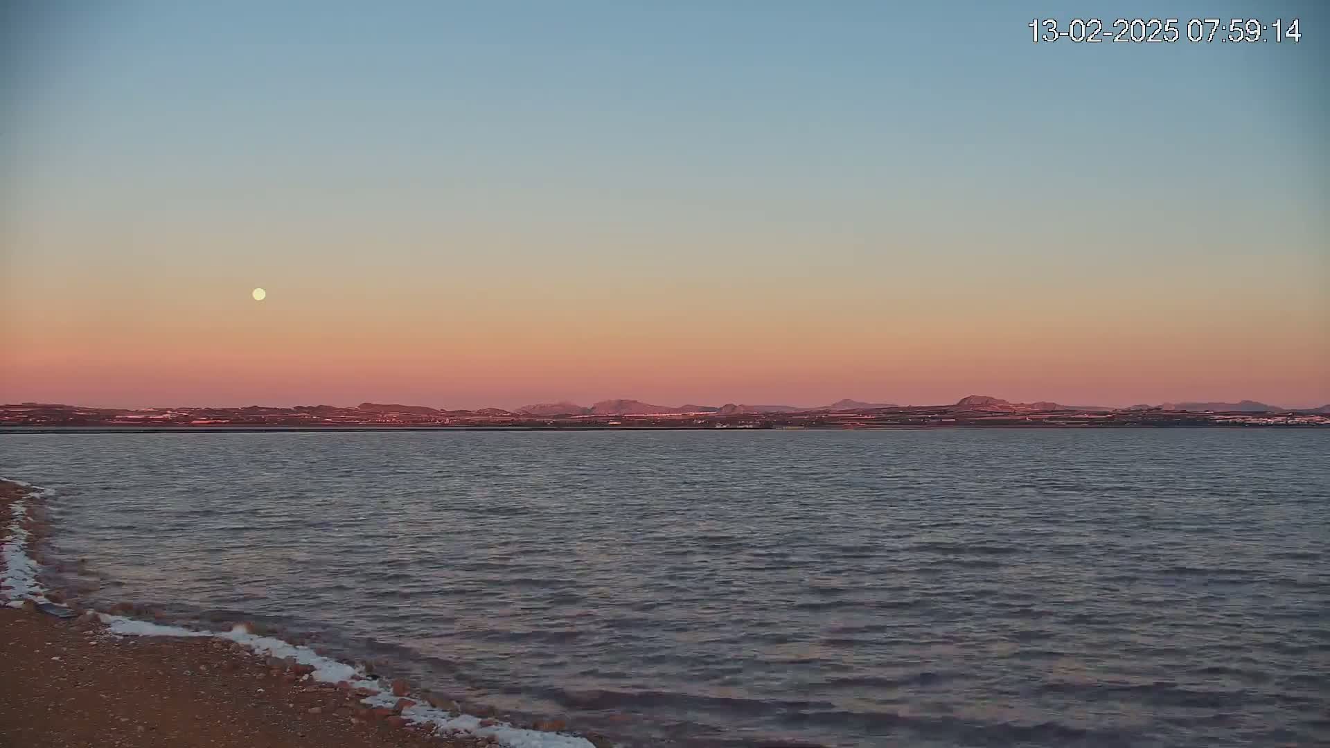 A calm body of water with a gently sloping sandy shore shows a pinkish sunset with a visible moon in the sky above a distant landmass.