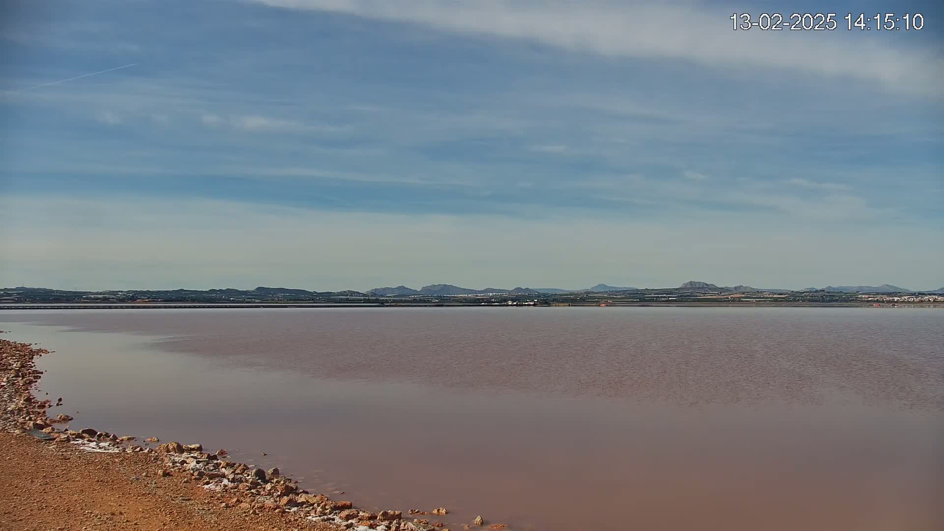 A calm, pinkish-hued body of water stretches towards a distant, low-lying mountain range under a mostly clear, light blue sky.