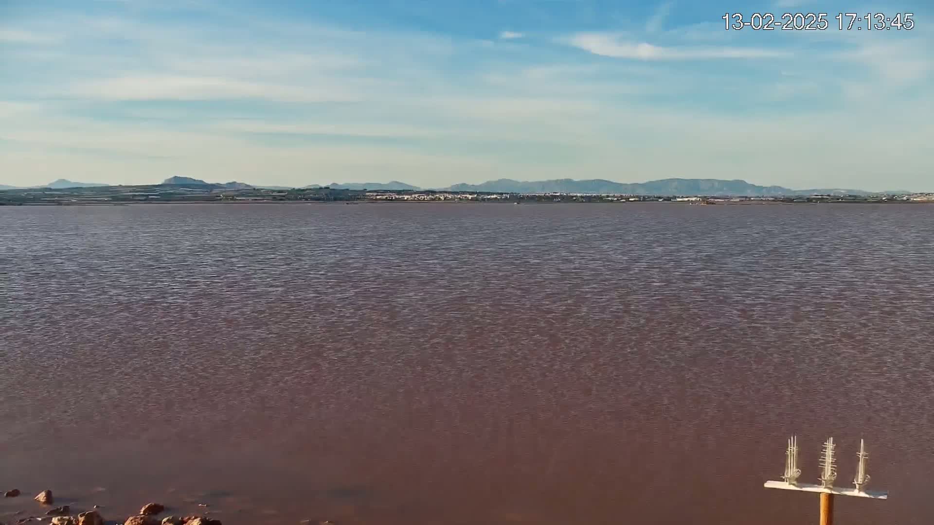 A calm, pink-hued body of water stretches to a distant line of low hills under a mostly clear sky.