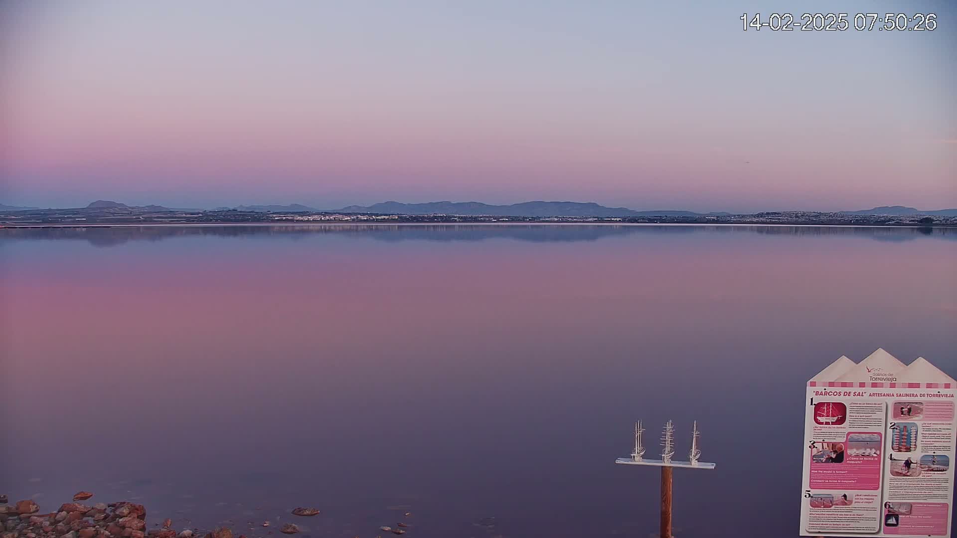 A calm, pink-hued body of water reflects a distant mountain range under a pale, pastel sky at dawn.