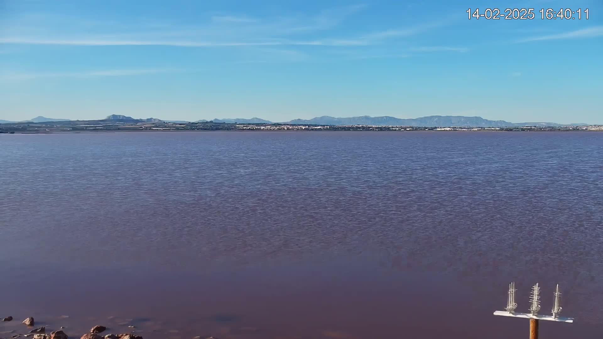 A calm, purplish-pink body of water stretches to a distant shoreline under a clear blue sky, with mountains visible on the horizon.