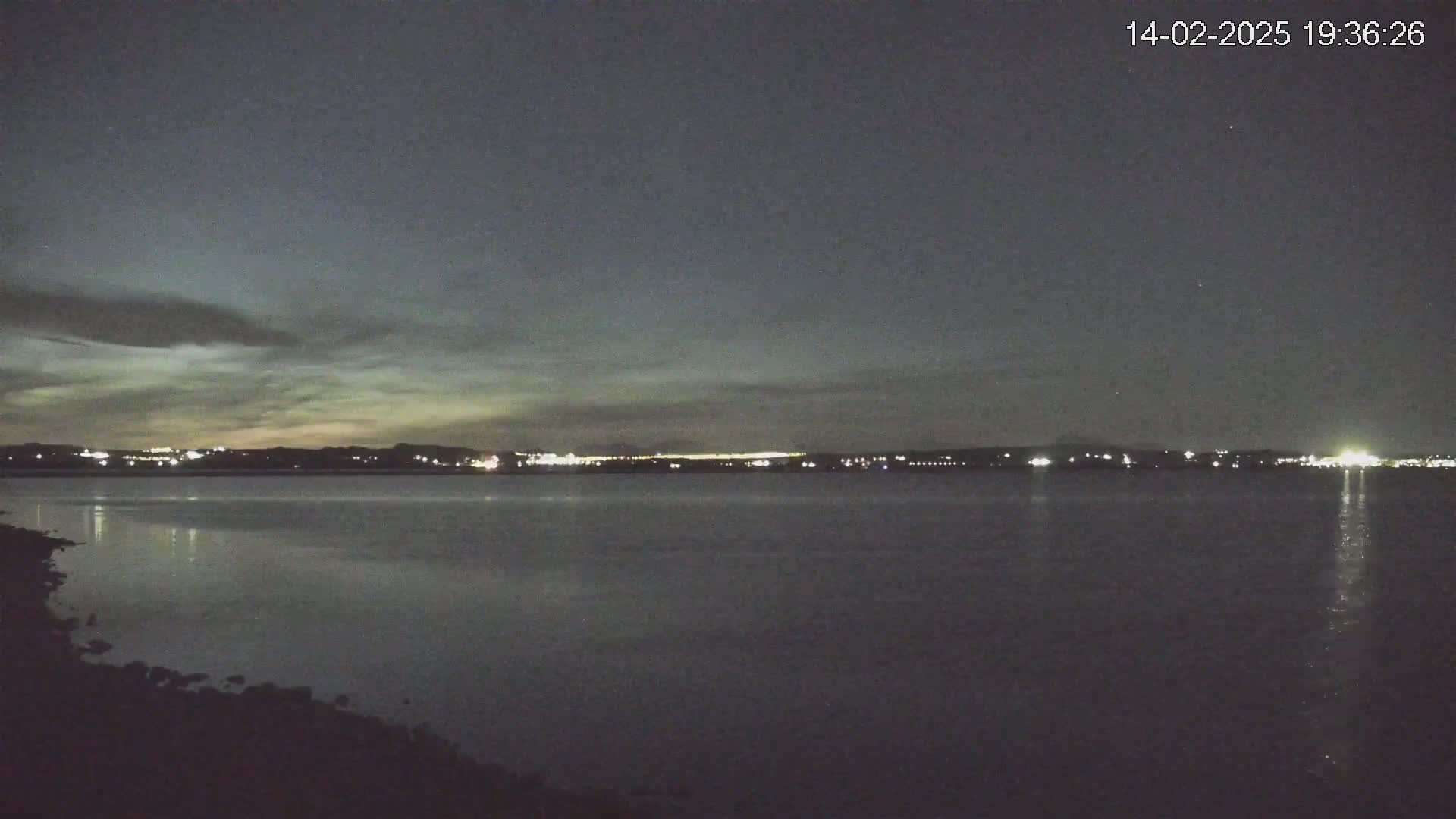 A dark, calm body of water reflects the lights of a distant city skyline under a mostly cloudy night sky.