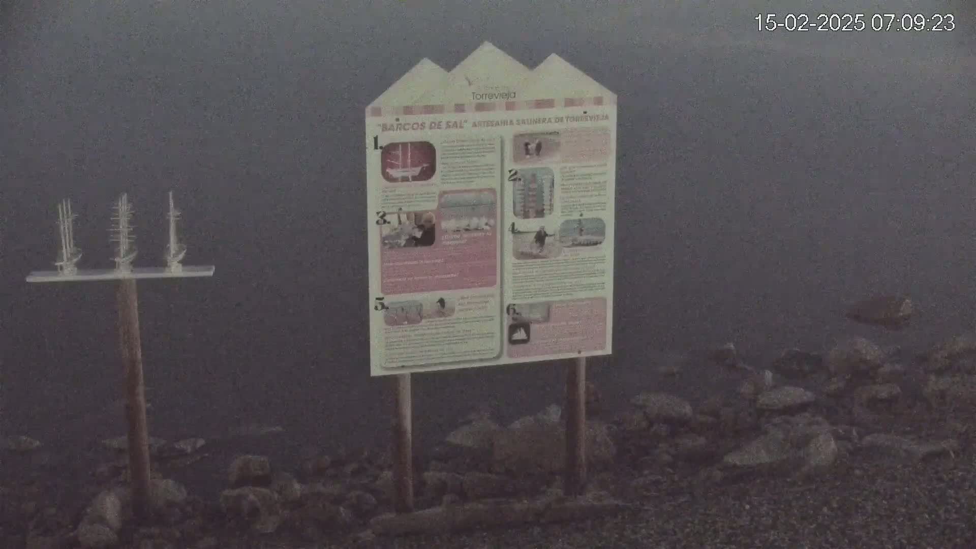 A foggy shoreline displays a wooden post with small model sailboats and an informational sign.