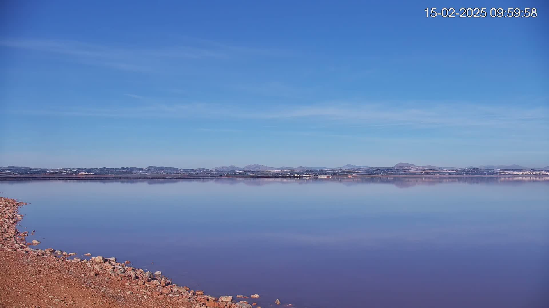 A calm, purplish lake reflects a distant, low mountain range under a clear blue sky.