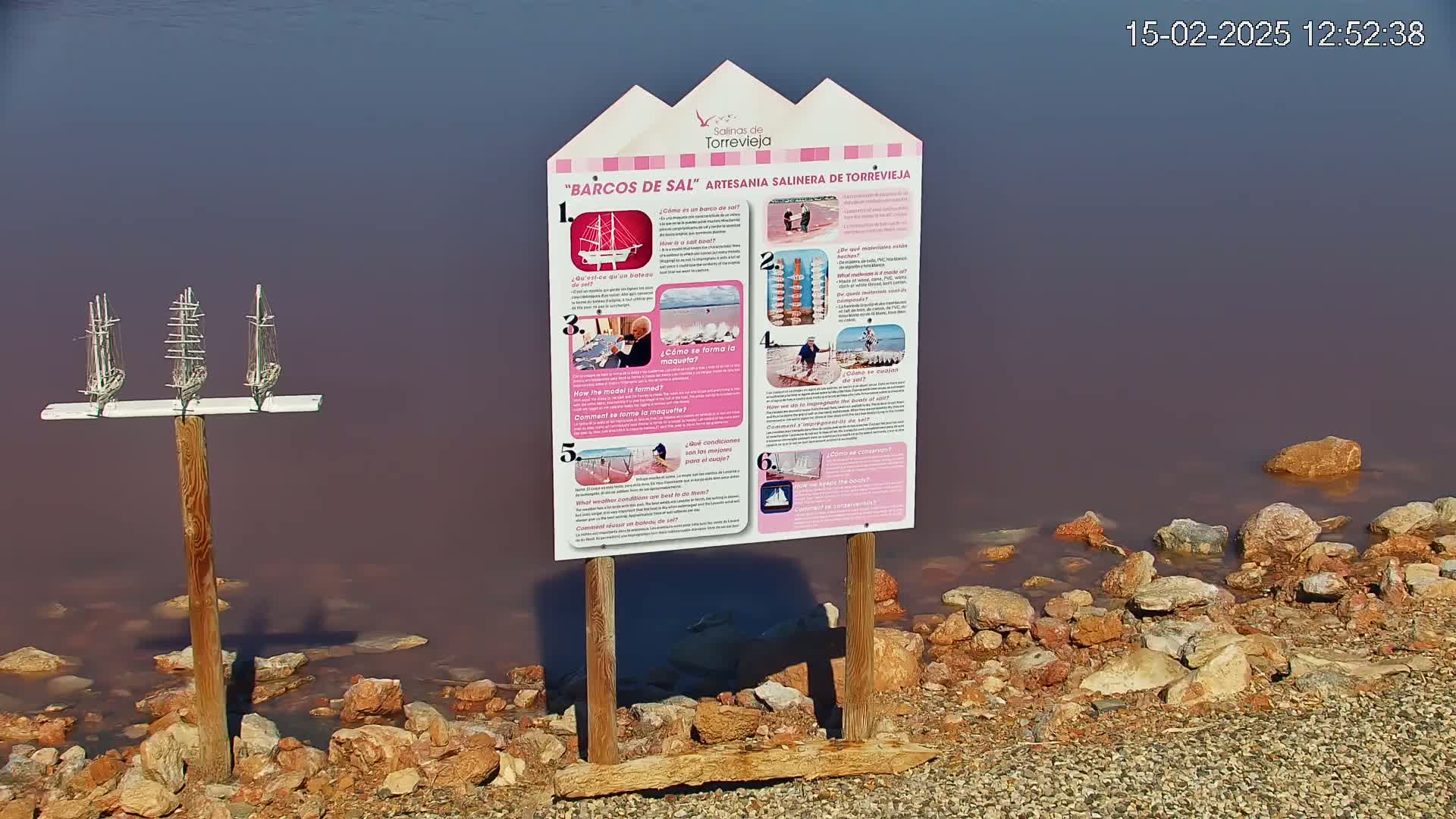 Three small model sailboats are mounted on a post near a pink-hued body of water with rocks along the shore.