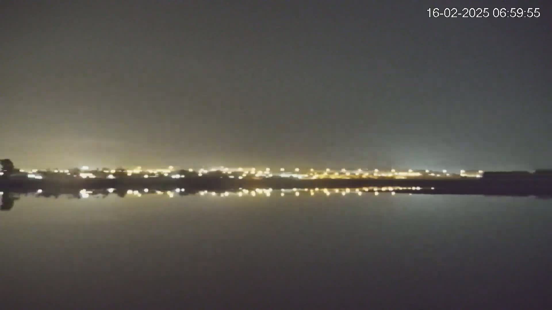 A calm body of water reflects the lights of a distant city at night under a dark sky.