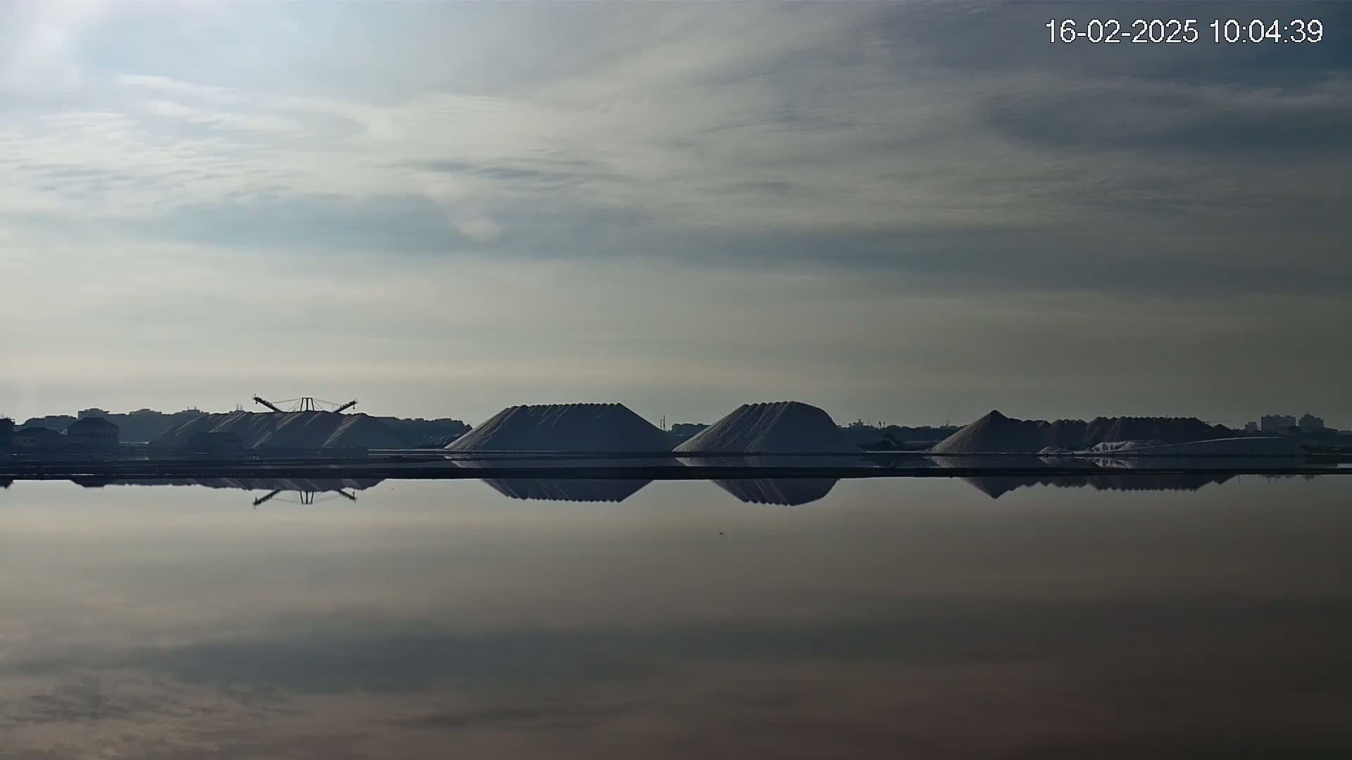 Several large piles of material, possibly salt, are situated near a calm body of water under a cloudy sky.