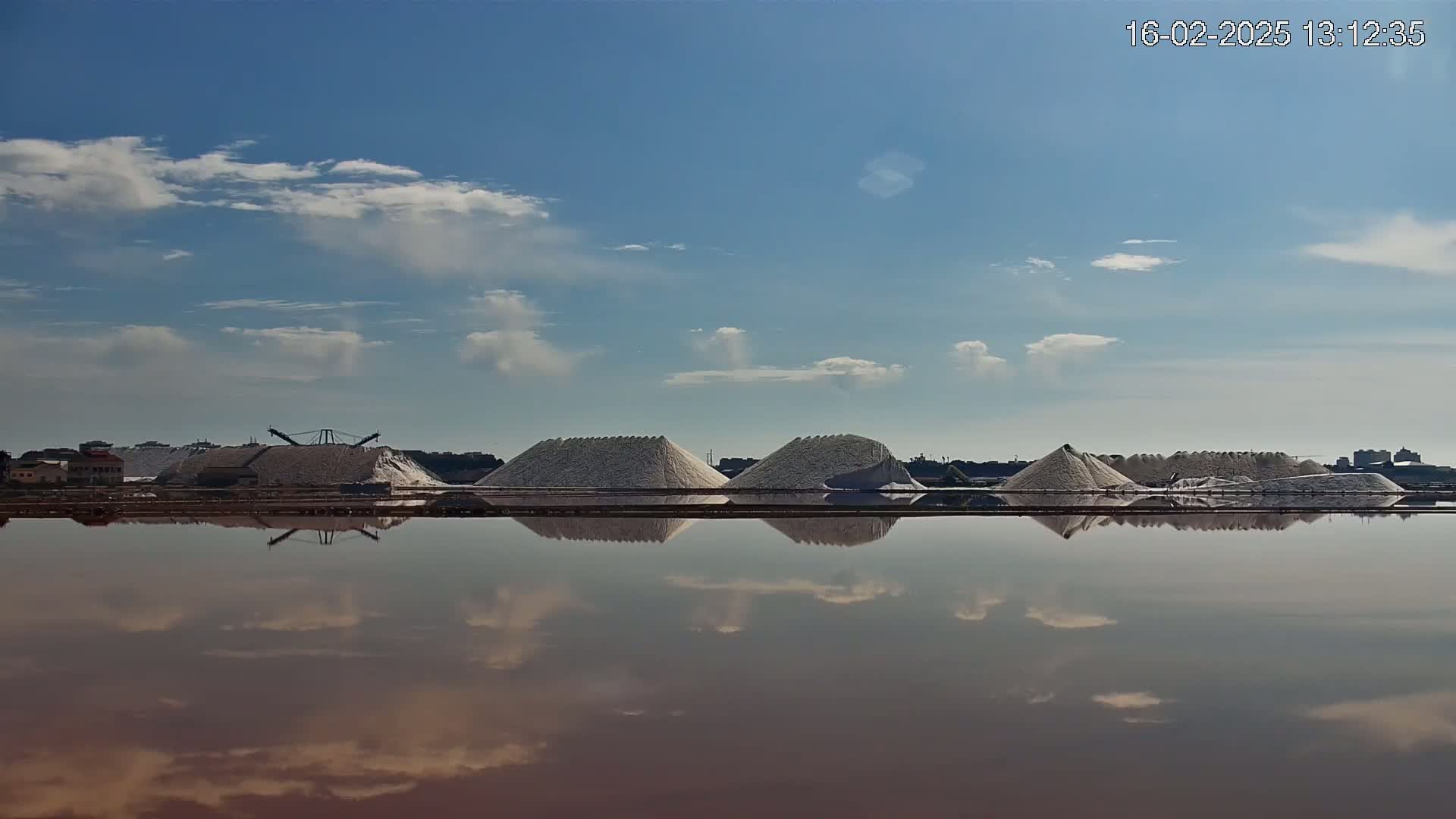 Several large piles of white salt are reflected in a calm body of reddish water under a partly cloudy blue sky.