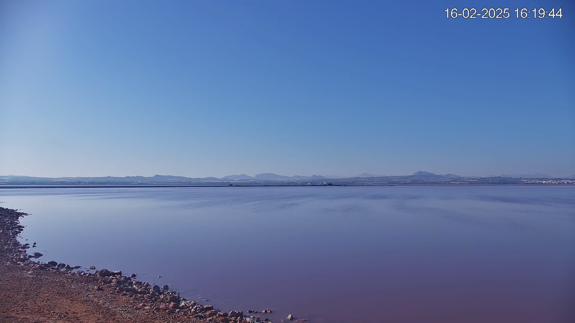 A calm, purplish-pink body of water stretches to a distant mountain range under a clear blue sky.