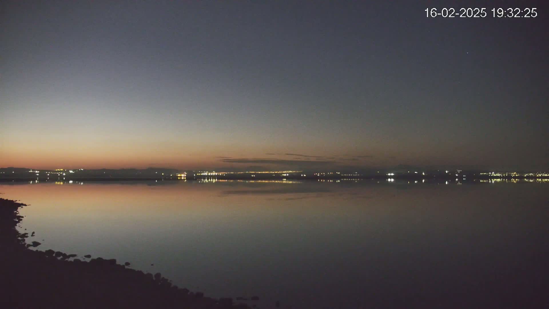 A calm body of water reflects the lights of a distant city skyline under a twilight sky.