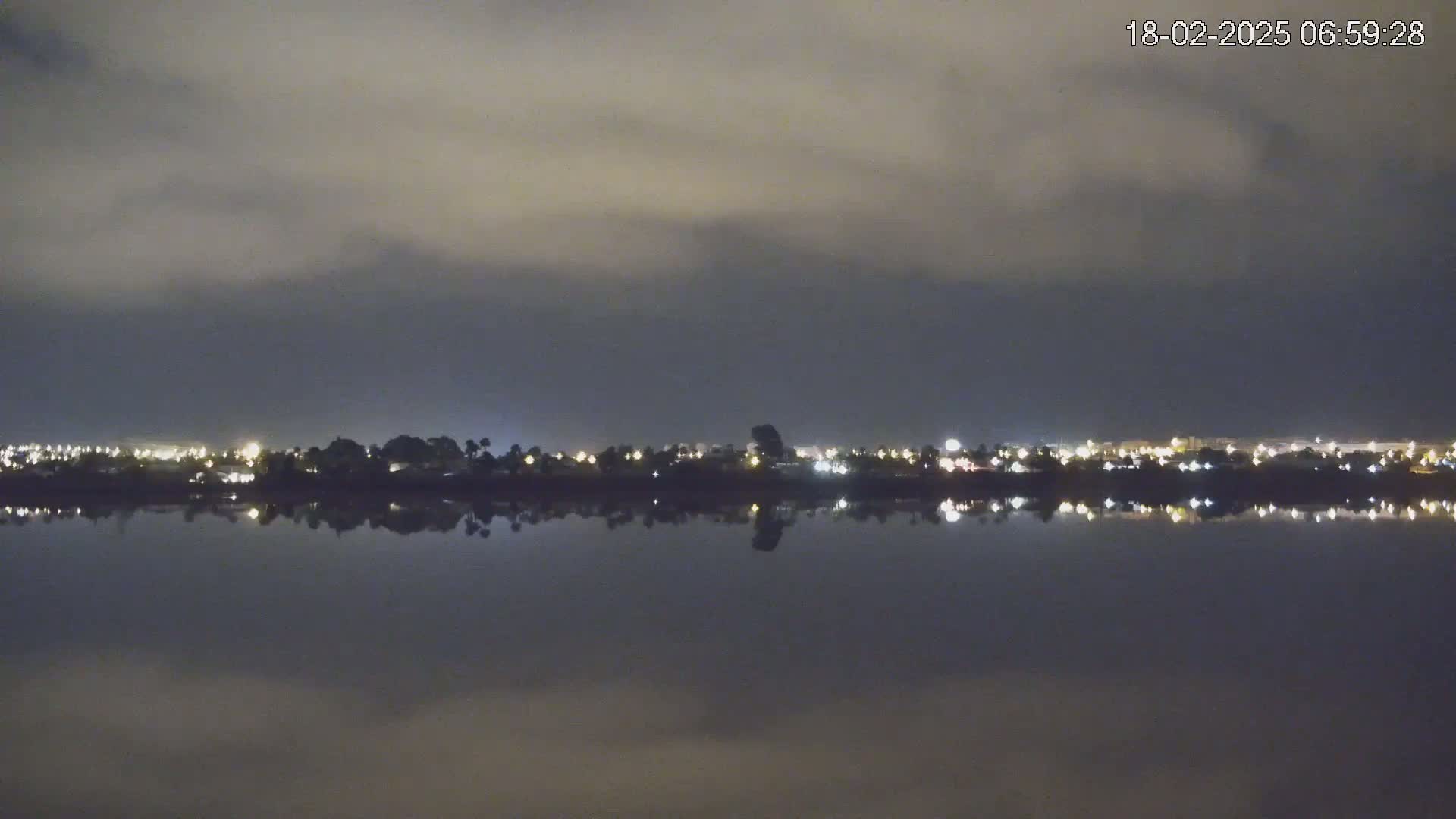 A calm body of water reflects the lights of a distant city under a cloudy night sky.