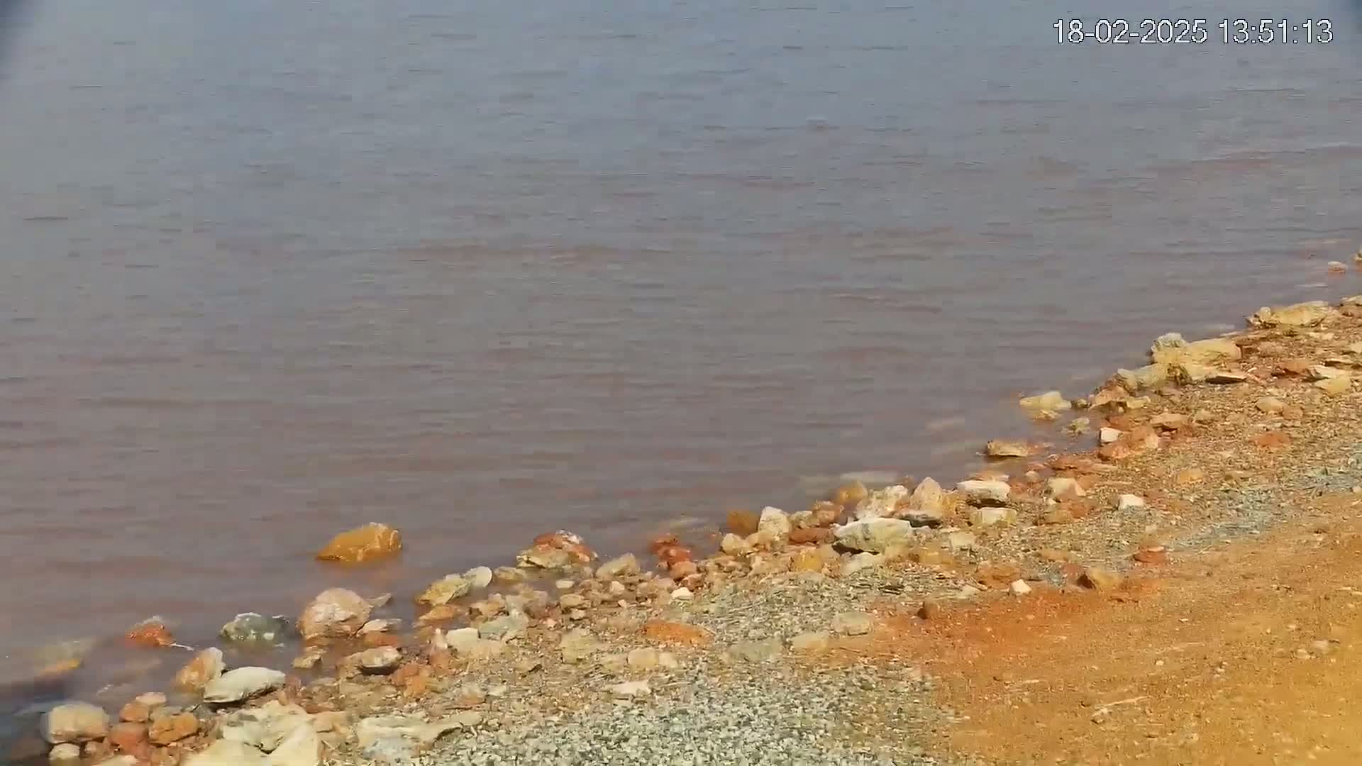 Calm, reddish-brown water laps a rocky shore composed of various sized stones and reddish-brown sand.