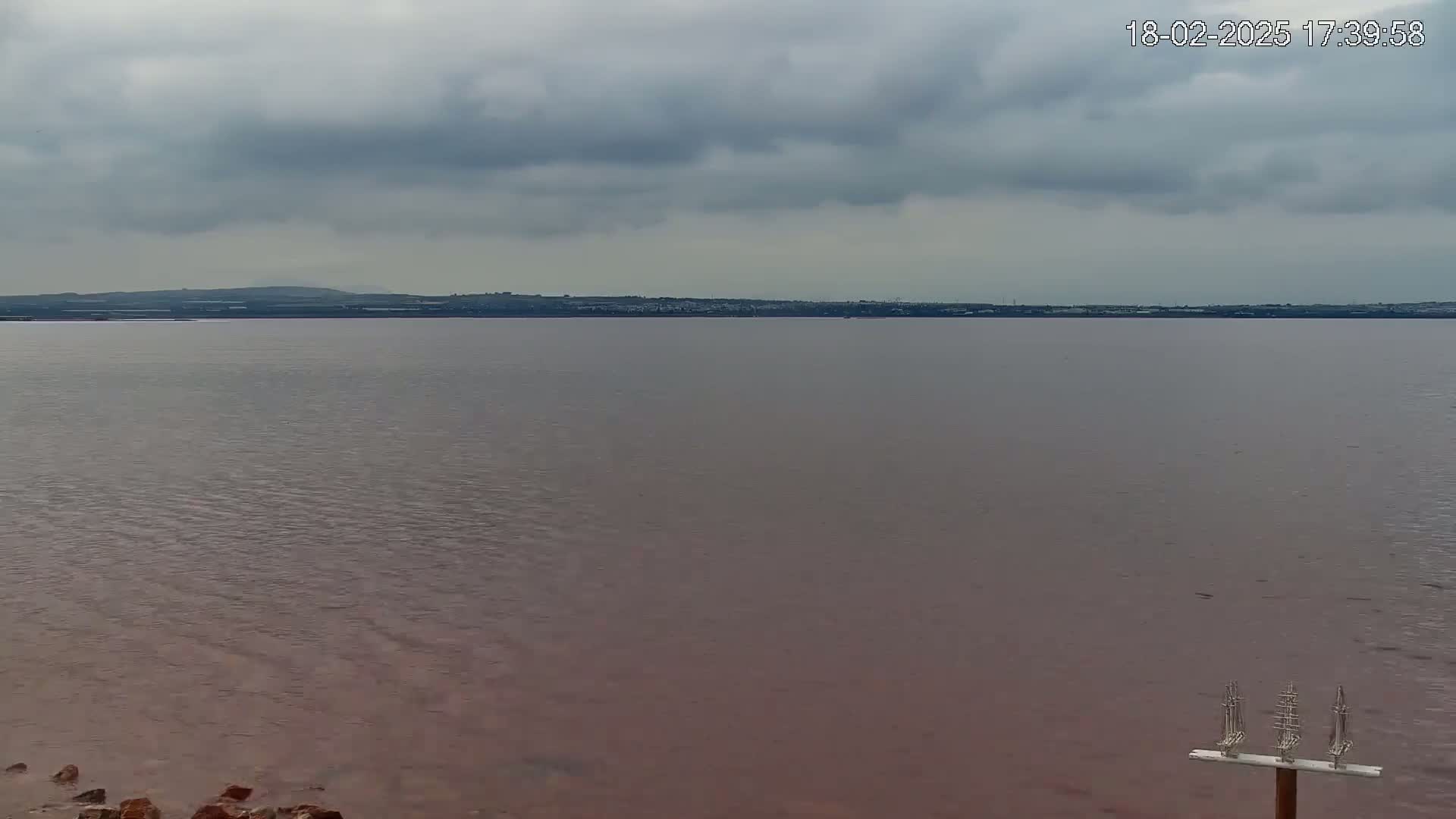 A calm, reddish-brown body of water stretches to a distant shoreline under a cloudy sky.