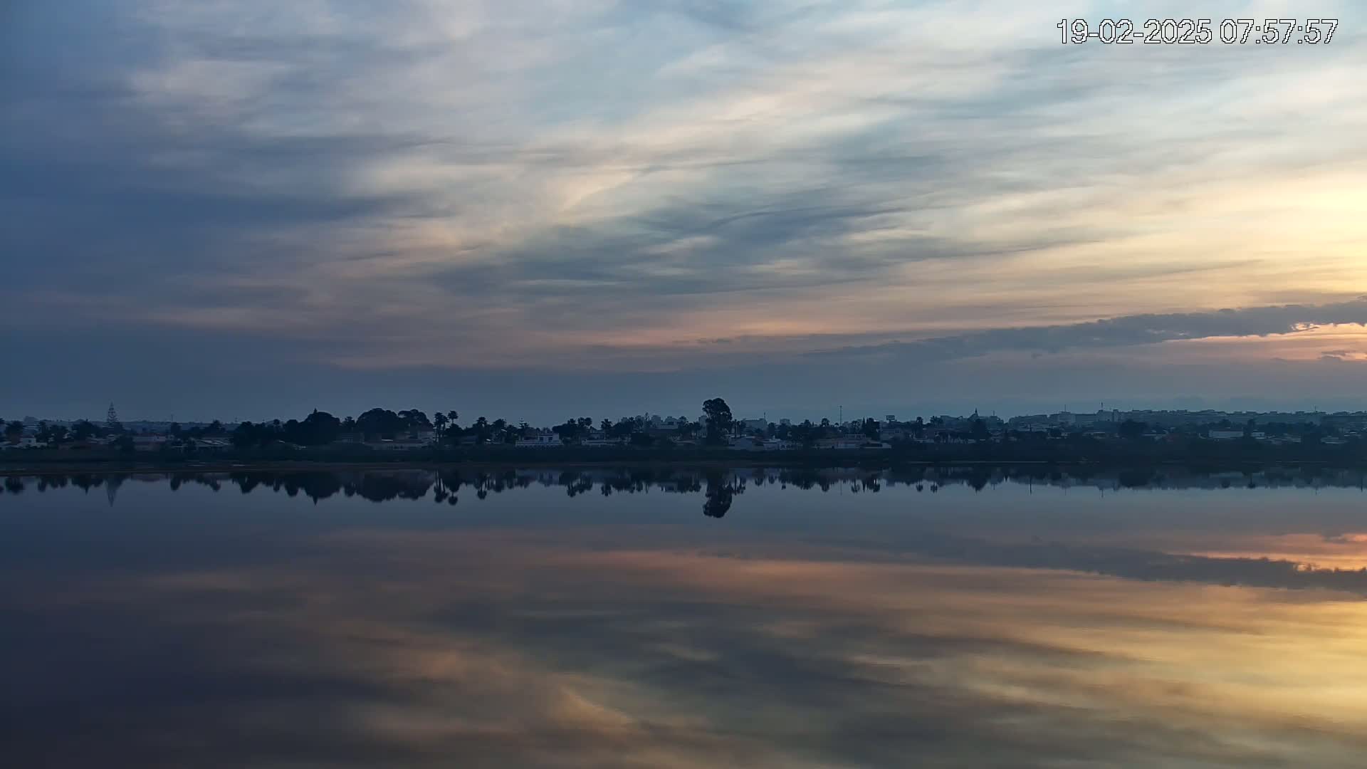 A calm body of water reflects a muted sunrise sky and a distant shoreline of trees and buildings.
