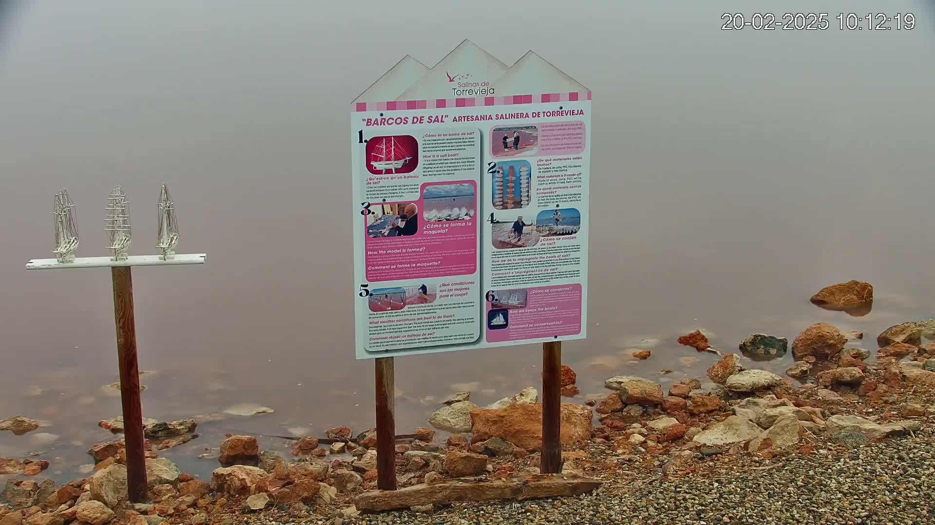 A foggy scene shows three small model sailboats on a post near a pink-tinged body of water, with a rocky shore and an informational sign about salt boats.