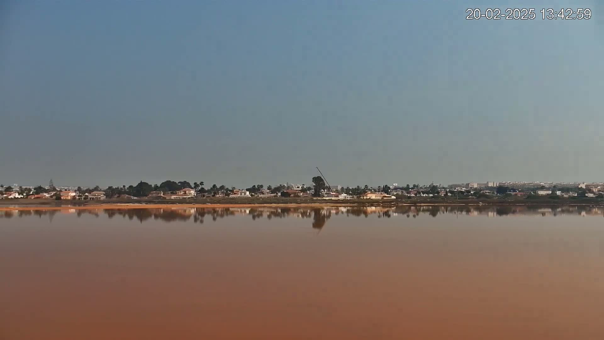 A calm, reddish-brown body of water reflects a clear sky and a line of houses and trees on the far shore.